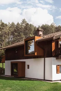 Exterior view of a contemporary mountain house featuring a large cantilevered Corten steel box with black-framed windows, set against a dense pine forest under a blue sky.