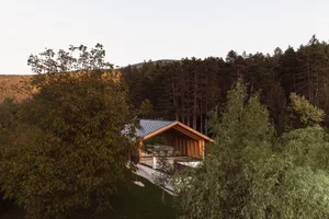 Modern wooden cabin with extensive glass walls situated on a forested hillside overlooking mountains at dusk, with a woman standing on the terrace.