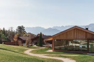Modern wooden alpine house with large glass windows overlooking a mountain range and autumnal landscape.