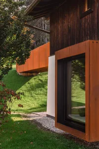 Modern wooden cabin exterior featuring a large cantilevered Corten steel window seat overlooking a green grassy slope and forest trees.