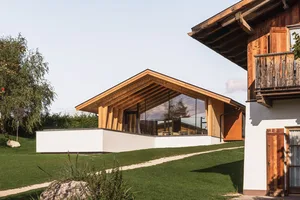 Modern alpine mountain house featuring large glass walls, exposed wooden beams, white concrete base and traditional wooden balcony under a clear sky.