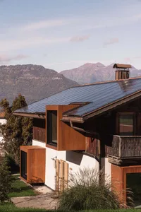 Modern mountain house with white walls, weathered corten steel architectural extensions, large solar panels on the roof, wooden balcony, and panoramic view of forested mountains in the background.