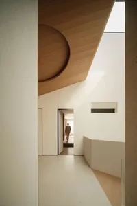 Modern minimalist hallway featuring a large curved oak wood ceiling detail, white walls, and a person walking toward a brightly lit room in the distance.
