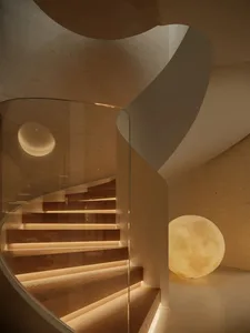 Modern curved wooden staircase with integrated LED lighting, glass railing, textured concrete walls, and two large spherical moon lamps providing soft illumination.