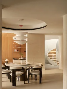 Modern dining room featuring a round marble table, dark wood upholstered chairs, a large multi-tiered pendant light, travertine walls, and a dramatic white spiral staircase in the background.