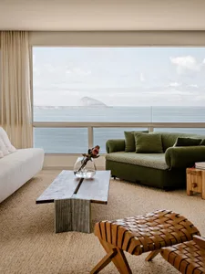 Modern coastal living room with green boucle sofa, white linen sofa, wooden coffee table, and large panoramic ocean window overlooking the sea and distant island.