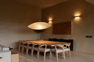 Modern minimalist dining room featuring a long solid oak dining table, eight matching oak dining chairs with light upholstery, a large sculptural pendant lamp, dark wood sideboard, and textured plaster walls in warm neutral tones.