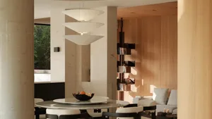 Modern dining room featuring a round travertine table, sculptural three-tier pendant light, light wood paneling, and built-in bookshelf in the background.
