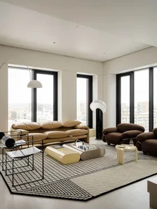 Modern minimalist living room featuring a tan leather sofa, brown boucle armchairs, black and white geometric rug, metallic coffee table, and large arched floor lamps with city views through floor-to-ceiling windows.