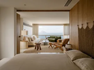 Modern coastal bedroom with beige linen bedding, woven teak headboard, and panoramic ocean view through the adjacent living room.