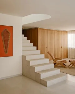 Modern interior featuring a floating terrazzo staircase, light wood built-in storage, woven rattan lounge chair and ottoman, and an orange artwork with woven textile on a white wall.