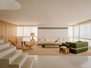 Modern living room featuring light oak wall paneling, a long white sofa, a curved green bouclé sofa, wooden lounge chairs, and a large jute rug under bright natural light.