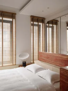 Modern minimalist bedroom with a large red travertine stone headboard, white bedding, wooden Venetian blinds, and a sculptural white table lamp.