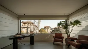 Modern minimalist home office featuring two cognac leather armchairs, a dark wood oval desk, large picture window with city view, and a tall potted plant.