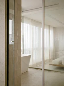 Modern minimalist bathroom interior with beige travertine tiled walls, white freestanding bathtub, sheer white curtains, and a floating bed visible through glass partition.
