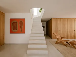 Modern minimalist entrance hall with a wide travertine stone staircase, large orange woven artwork, light wood cabinetry, and a woven leather lounge chair under a circular skylight.