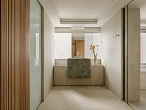 Modern minimalist bathroom with a large monolithic dark stone vanity, travertine walls and floor, vertical wood slat accent wall, and a vase of orange flowers.