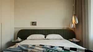 Modern minimalist bedroom with a large green velvet upholstered bed, white linens, a colorful checkered throw blanket, two round side tables, and three sculptural metallic pendant lights hanging above the right side.