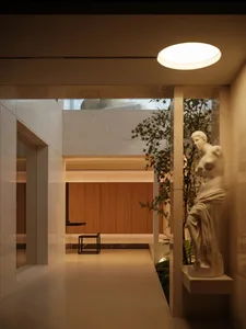 Modern minimalist hallway featuring travertine walls and floors, warm wood paneling, a classical white marble Venus statue, and lush indoor plants under a circular ceiling light.