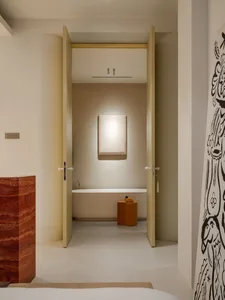 View into a minimalist bathroom with a freestanding white bathtub, a glowing wall sconce, an ochre terrazzo stool, and a red marble block in the foreground.