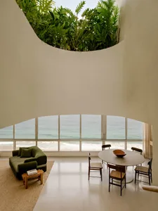 Modern open-plan living and dining room with curved white walls, green velvet sofa, round dining table, and large ocean view windows overlooking the sea and beach.