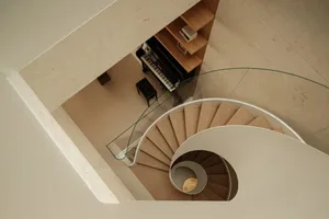 Top-down view of a modern spiral staircase made of light oak with white curved balustrade, glass railing, and a black upright piano nestled in a built-in wooden shelving nook.