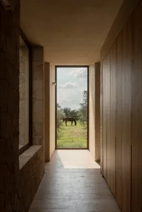 Narrow hallway with rough stone wall on the left, light wood paneling on the right, concrete floor, and a large floor-to-ceiling window at the end revealing a horse grazing in a green pasture under a blue sky.