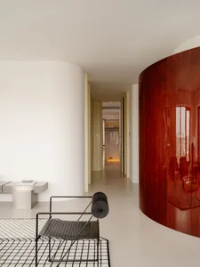 Modern minimalist hallway featuring a large curved wardrobe in glossy rosewood, black metal chair on a black and white rug, and a view into a walk-in closet with hanging clothes.