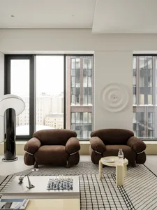 Modern minimalist living room featuring two brown boucle armchairs, a black and white grid-patterned rug, a low white coffee table with a chess set, and large floor-to-ceiling windows overlooking city buildings.