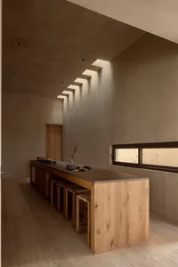 Modern minimalist kitchen featuring a long solid oak island with concrete countertop, four woven stools, raw concrete walls and ceiling, and a series of stepped skylights casting dramatic light patterns.