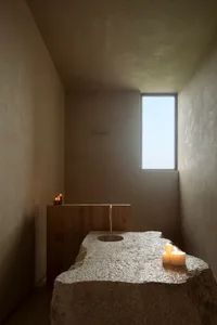 Minimalist bathroom featuring a massive rough-hewn limestone slab with integrated round basin, wooden vanity, brass faucet, and lit candles under a tall vertical window.