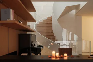Modern interior featuring a sweeping curved oak staircase, black grand piano, built-in wooden bookshelves, and lit candles on a dark table with autumn leaves.