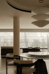 Modern dining room featuring a large round travertine table, curved upholstered chairs, a cylindrical concrete column, and a sculptural three-tiered pendant light with large windows overlooking bamboo in the background.