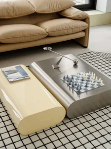 Modern living room scene featuring a tan leather sofa, a glossy cream curved coffee table, and a brushed stainless steel coffee table with a chessboard and a sculptural metal lamp on a black and white grid rug.