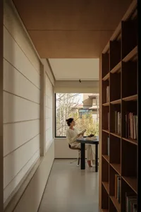 Woman reading in a minimalist oak wood reading nook with tall built-in bookshelves and large window overlooking trees