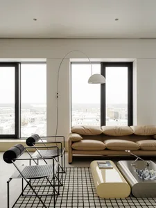 Modern minimalist living room with beige leather sofa, two black wire chairs on a black and white grid rug, glossy curved coffee tables, and large windows showing a snowy city view.