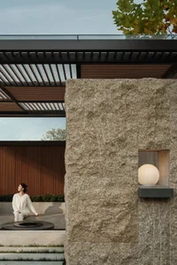 Modern outdoor courtyard featuring a large travertine stone wall with a recessed glowing sphere lamp, wooden pergola, circular fire pit, and a woman in white sitting on stone steps.