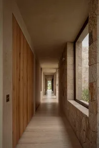 Long minimalist corridor featuring light wood wall paneling on the left and textured natural stone masonry on the right, with large windows allowing daylight to illuminate the concrete floor.