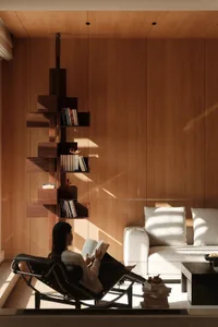 Woman reading in a black leather chaise lounge beside a sculptural walnut bookshelf in a sunlit modern living room with a light-colored sofa.