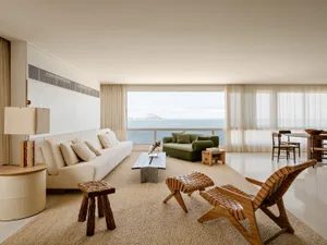 Modern coastal living room featuring a long white sofa, green velvet sofa, wooden woven leather lounge chair, and large floor-to-ceiling windows overlooking the ocean.