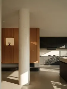 Modern minimalist kitchen interior featuring a prominent cylindrical white marble column, warm oak wood cabinetry, dark grey kitchen units, and strong natural sunlight creating sharp shadows on the floor.