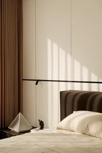 Modern minimalist bedroom corner featuring a dark taupe ribbed fabric headboard, cream bedding, a black floating shelf, and a brown curtain with strong morning sunlight creating striped shadows on the wall.