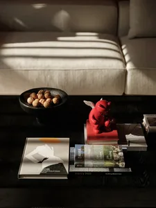 Close-up of a dark walnut coffee table in a living room featuring a red winged figurine, a bowl of walnuts, and stacked design books including Dior and Kerry Hill.