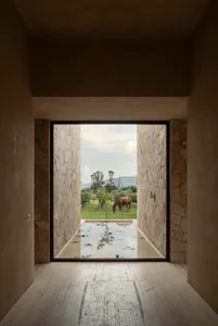 View from inside a minimalist concrete and stone room looking out through a large opening to a shallow reflecting pool, stone walls, green pasture, and a brown horse grazing with distant mountains.