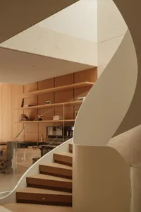 Modern curved oak staircase with white curved wall detail, floating wooden shelves, glass railing, and a grand piano visible in the background of a minimalist living room.