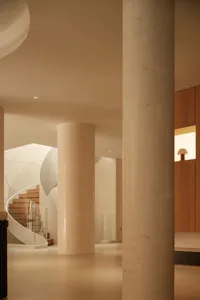 Modern interior featuring large cylindrical travertine columns, a sculptural curved wooden staircase with glass railing, and warm ambient lighting.