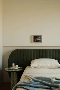 Modern bedroom with curved dark green velvet headboard, white linen bedding, blue textured throw blanket, and a unique green marble side table with candles and books against a two-tone wall with framed art.