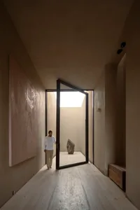 Minimalist concrete hallway with large abstract wall art, black-framed pivot door opening to a sunlit courtyard containing a large natural stone sculpture.