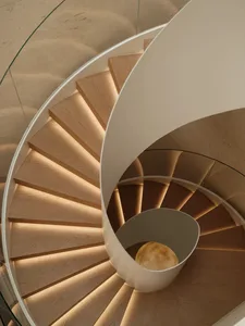 Top-down view of a modern spiral staircase made of light oak with integrated warm LED lighting under each step, featuring a white curved central structure and glass railing.