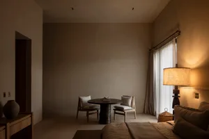 Modern minimalist bedroom interior featuring a dark oak round table with two upholstered chairs, a tall woven floor lamp casting warm light, neutral beige walls, and a bed with textured linen bedding.